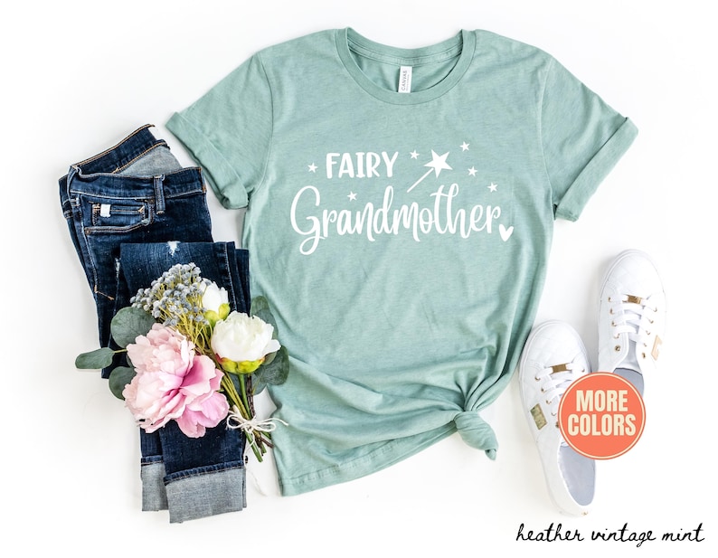 K&ouml;nnte beinhalten: Ein heather vintage mintfarbenes T-Shirt mit wei&szlig;em Schriftzug Fairy Grandmother, erg&auml;nzt durch eine Grafik eines Zauberstabs und Sternen. Das T-Shirt ist kombiniert mit einer dunkelblauen Jeans, einem Strau&szlig; rosa und wei&szlig;er Blumen und wei&szlig;en Turnschuhen.