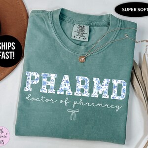 Puede incluir: Una camiseta azul turquesa con un estampado floral blanco que dice "PHARMD doctor of pharmacy".