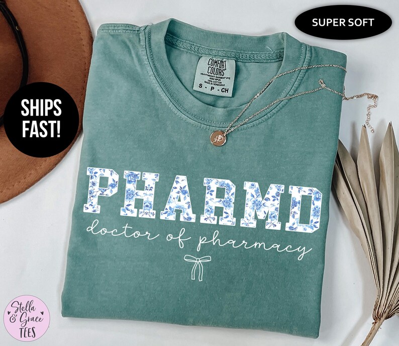 Puede incluir: Una camiseta azul turquesa con un estampado floral blanco que dice "PHARMD doctor of pharmacy".