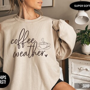 May include: Beige sweatshirt with the words "coffee weather" in a handwritten font, along with a coffee cup graphic. The sweatshirt is a neutral color and has long sleeves. The text is in a dark brown color.