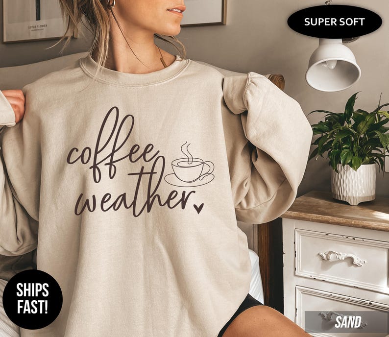 May include: Beige sweatshirt with the words "coffee weather" in a handwritten font, along with a coffee cup graphic. The sweatshirt is a neutral color and has long sleeves. The text is in a dark brown color.