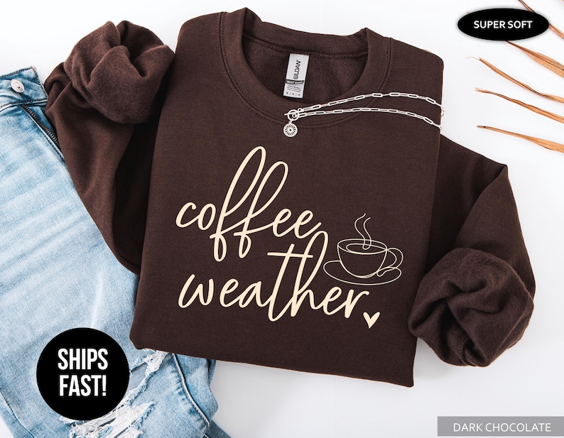 May include: Dark chocolate-colored sweatshirt with the words "coffee weather" in a cursive font, along with a coffee cup graphic. A silver necklace rests on the sweatshirt, which is folded next to a pair of blue jeans.