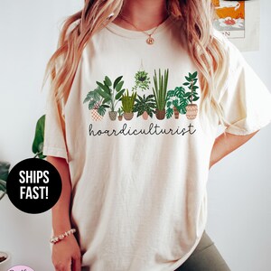 Op de afbeelding: Ivoor kleurig T-shirt met een afbeelding van verschillende potplanten en het woord "hoardiculturist". Het shirt is voorzien van de labels "Super Soft" en "Snel verzonden!"