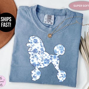 May include: A blue Comfort Colors t-shirt featuring a poodle silhouette filled with a blue floral pattern. The shirt has a crew neck and short sleeves. The text "SUPER SOFT" is above the shirt. A gold necklace is on the shirt.