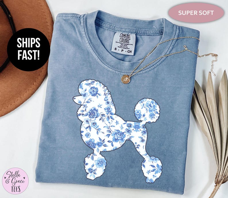 May include: A blue Comfort Colors t-shirt featuring a poodle silhouette filled with a blue floral pattern. The shirt has a crew neck and short sleeves. The text "SUPER SOFT" is above the shirt. A gold necklace is on the shirt.