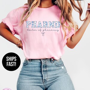 Puede incluir: Camiseta rosa con el texto "PHARMD doctor of pharmacy" en letras azules con contorno blanco. La camiseta tiene un patr&oacute;n floral en azul y blanco.