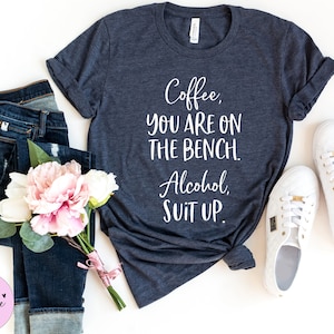 May include: A dark blue t-shirt with white text that reads "Coffee, you are on the bench. Alcohol, suit up."