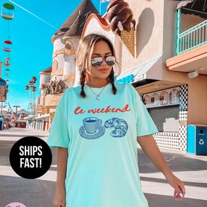 May include: A light turquoise t-shirt with the words "Le weekend" in red script above a blue illustration of a coffee cup and croissant. The shirt is labeled "Super Soft" and "Ships Fast!"
