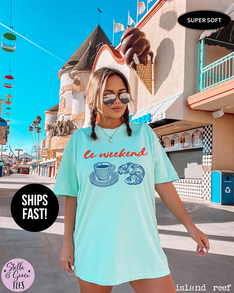 May include: A light turquoise t-shirt with the words "Le weekend" in red script above a blue illustration of a coffee cup and croissant. The shirt is labeled "Super Soft" and "Ships Fast!"