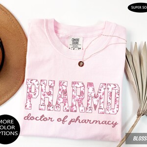 May include: A light pink t-shirt featuring the text PHARMD in a floral pattern and the words doctor of pharmacy in a cursive font. The shirt has a Comfort Colors label at the neck and is displayed with a necklace, a hat, and a dried palm leaf.