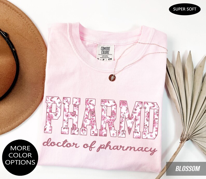 May include: A light pink t-shirt featuring the text PHARMD in a floral pattern and the words doctor of pharmacy in a cursive font. The shirt has a Comfort Colors label at the neck and is displayed with a necklace, a hat, and a dried palm leaf.