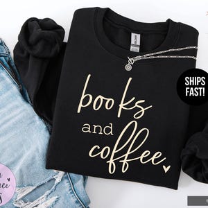 May include: Black sweatshirt with the words "books and coffee" in a cursive font. A silver necklace is draped across the top. The sweatshirt is next to a pair of blue jeans.