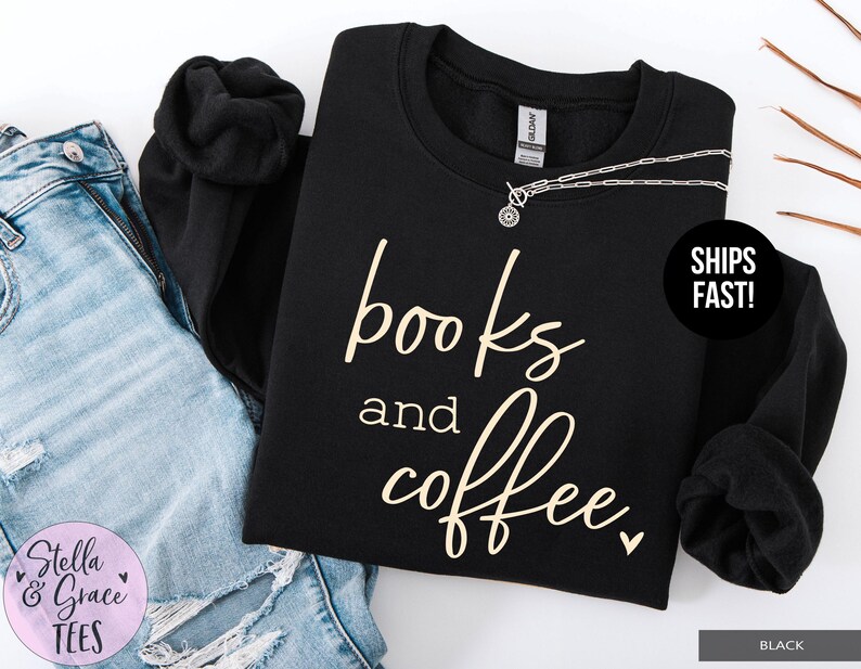 May include: Black sweatshirt with the words "books and coffee" in a cursive font. A silver necklace is draped across the top. The sweatshirt is next to a pair of blue jeans.