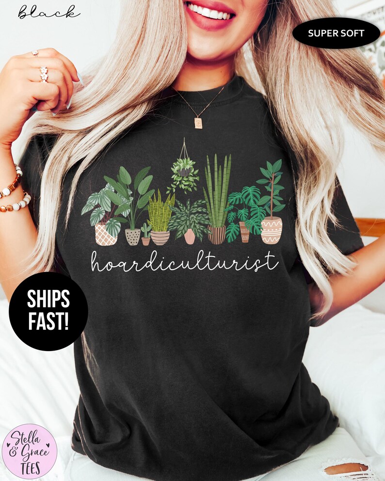 Op de afbeelding: Zwart T-shirt met een botanisch ontwerp met diverse potplanten en het woord "hoardiculturist" in witte letters. Het shirt is gelabeld met "Super Soft" en heeft een ronde hals. De merknaam "Stella & Grace Tees" is ook zichtbaar.