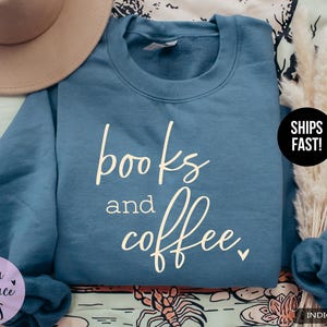 May include: Indigo blue sweatshirt with the words "books and coffee" in a cursive font. The sweatshirt is a crew neck style. A beige hat and dried flowers are in the background.