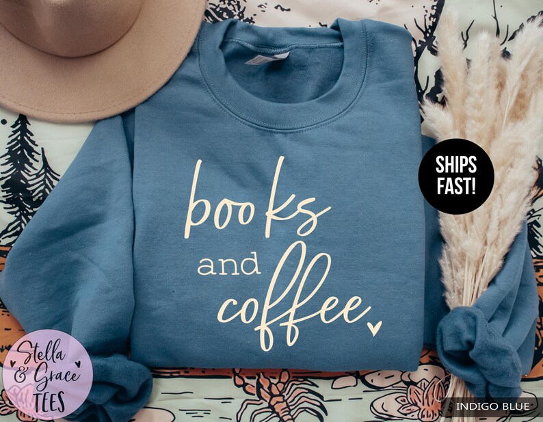 May include: Indigo blue sweatshirt with the words "books and coffee" in a cursive font. The sweatshirt is a crew neck style. A beige hat and dried flowers are in the background.