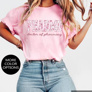 May include: A light pink short-sleeved t-shirt featuring the text PHARMD in a floral pattern with the words doctor of pharmacy printed underneath in a cursive font. The shirt includes black circular labels stating SUPER SOFT and MORE COLOR OPTIONS.
