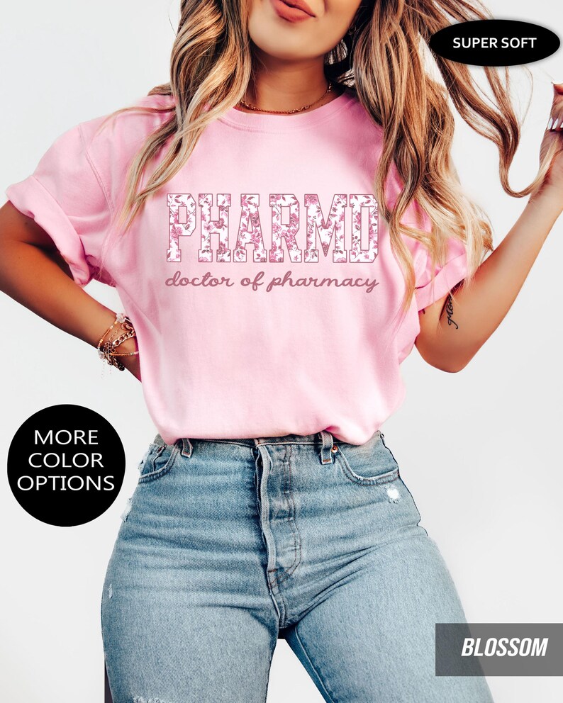 May include: A light pink short-sleeved t-shirt featuring the text PHARMD in a floral pattern with the words doctor of pharmacy printed underneath in a cursive font. The shirt includes black circular labels stating SUPER SOFT and MORE COLOR OPTIONS.