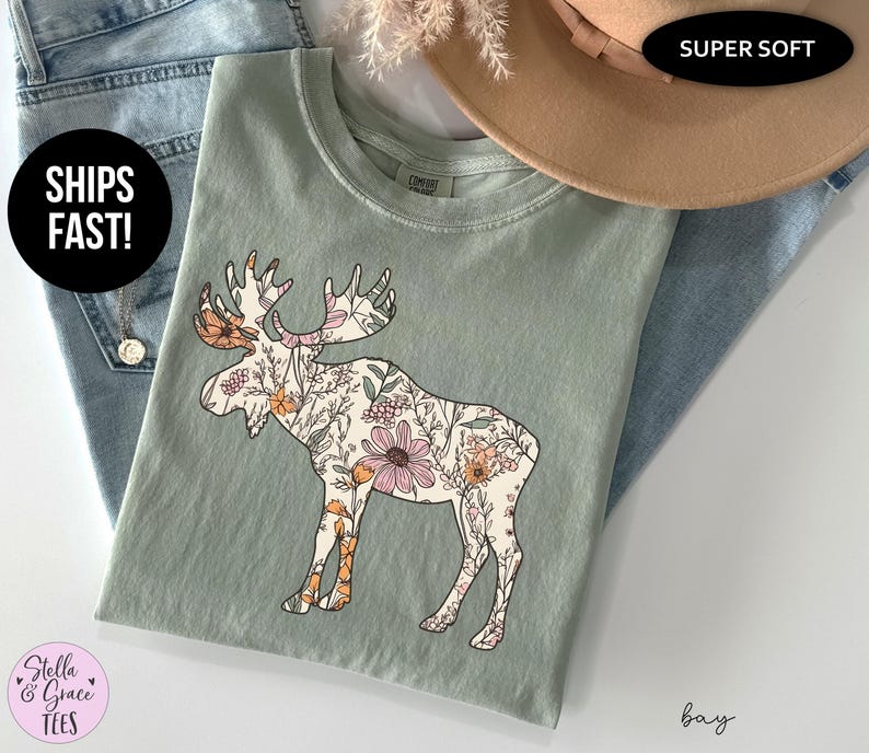May include: A sage green crew neck t-shirt featuring a large graphic of a moose filled with a detailed floral pattern in shades of pink, orange, and cream. The shirt has a ribbed collar and a relaxed fit, showcasing a nature-inspired design.