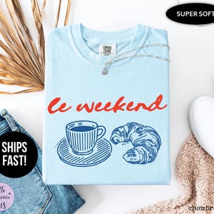 May include: Light blue t-shirt with the words "Le weekend" in red script. The shirt features a blue illustration of a coffee cup and croissant. A silver necklace is draped on the shirt. The shirt is labeled "Super Soft".