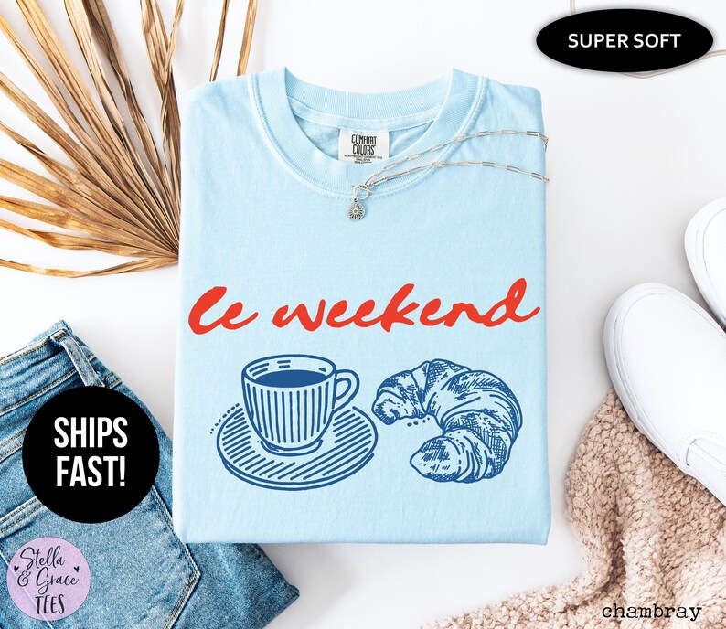 May include: Light blue t-shirt with the words "Le weekend" in red script. The shirt features a blue illustration of a coffee cup and croissant. A silver necklace is draped on the shirt. The shirt is labeled "Super Soft".