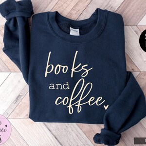 May include: Navy blue sweatshirt with the words "books and coffee" in a cursive font. The sweatshirt has a crew neck and long sleeves. The text is in a light beige color. The brand name "Stella & Grace Tees" is in a pink circle.