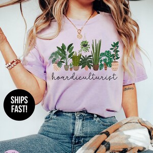 Op de afbeelding: Lichtpaars T-shirt met een afbeelding van verschillende potplanten en het woord "hoardiculturist" in script. Het shirt heeft ook de woorden "Super Soft" en "Ships Fast!"