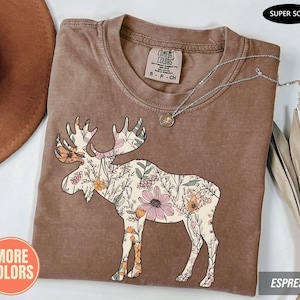May include: A brown short-sleeve t-shirt featuring a graphic of a moose filled with a floral pattern of pink, orange, and green flowers. The shirt has a Comfort Colors brand label at the neck, a crew neckline, and a silver-toned chain necklace resting on top.