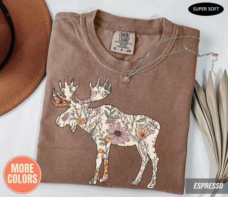 May include: A brown short-sleeve t-shirt featuring a graphic of a moose filled with a floral pattern of pink, orange, and green flowers. The shirt has a Comfort Colors brand label at the neck, a crew neckline, and a silver-toned chain necklace resting on top.