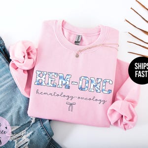 May include: Pink sweatshirt with the text "HEM-ONC hematology-oncology" in blue and white lettering with a small bow.