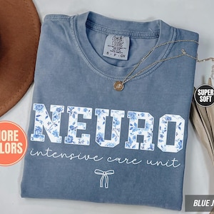 May include: A blue jean colored t-shirt with a crew neckline and a Comfort Colors label. The front features the word NEURO in block letters filled with a blue floral pattern, followed by the text intensive care unit and a small bow graphic.