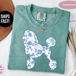 May include: A teal-colored t-shirt featuring a floral poodle design in shades of blue and white. The shirt has a crew neck and short sleeves. A silver necklace with a pendant is draped across the shirt. The text "SUPER SOFT" is in a pink oval.