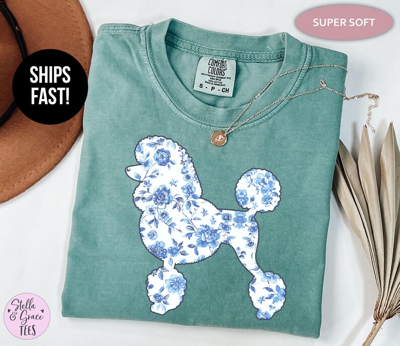 May include: A teal-colored t-shirt featuring a floral poodle design in shades of blue and white. The shirt has a crew neck and short sleeves. A silver necklace with a pendant is draped across the shirt. The text "SUPER SOFT" is in a pink oval.
