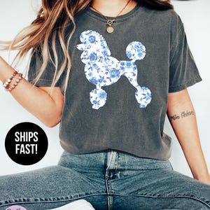 May include: Dark gray t-shirt featuring a poodle design with blue floral patterns. The shirt has a crew neck and short sleeves. The words "SUPER SOFT" are printed on the top right of the shirt. The words "SHIPS FAST!" are printed on a black circle on the bottom left of the shirt.