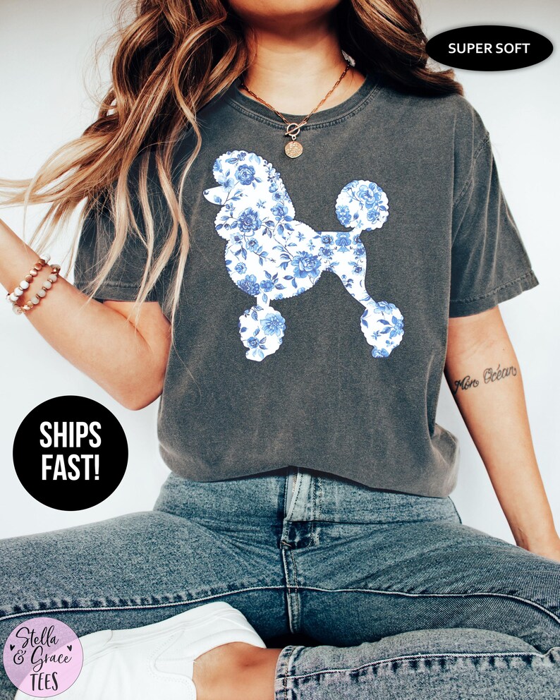 May include: Dark gray t-shirt featuring a poodle design with blue floral patterns. The shirt has a crew neck and short sleeves. The words "SUPER SOFT" are printed on the top right of the shirt. The words "SHIPS FAST!" are printed on a black circle on the bottom left of the shirt.