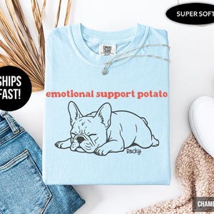 May include: A light blue t-shirt with the text "emotional support potato" above a black and white illustration of a French Bulldog. The shirt is folded and has a silver necklace on top.