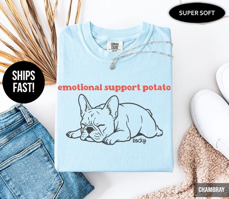 May include: A light blue t-shirt with the text "emotional support potato" above a black and white illustration of a French Bulldog. The shirt is folded and has a silver necklace on top.