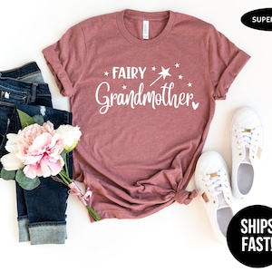 K&ouml;nnte beinhalten: Ein malvenfarbenes Kurzarm-T-Shirt mit dem wei&szlig;en Schriftzug "FAIRY Grandmother" und einer Grafik eines Zauberstabs mit Stern. Das T-Shirt wird zusammen mit einer dunkelblauen Jeans, einem Strau&szlig; rosa-wei&szlig;er Blumen und wei&szlig;en Turnschuhen gezeigt.