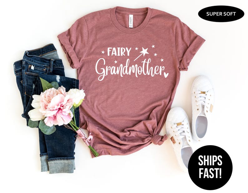 K&ouml;nnte beinhalten: Ein malvenfarbenes Kurzarm-T-Shirt mit dem wei&szlig;en Schriftzug "FAIRY Grandmother" und einer Grafik eines Zauberstabs mit Stern. Das T-Shirt wird zusammen mit einer dunkelblauen Jeans, einem Strau&szlig; rosa-wei&szlig;er Blumen und wei&szlig;en Turnschuhen gezeigt.