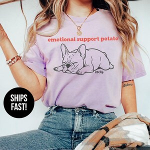 May include: A light purple t-shirt with the text "emotional support potato" above a cartoon illustration of a French Bulldog. The shirt also has the word "Rocky" and the words "Super Soft" printed on it.