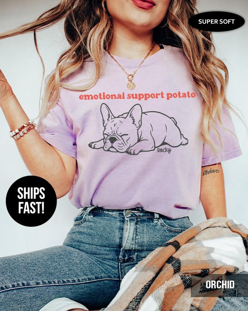 May include: A light purple t-shirt with the text "emotional support potato" above a cartoon illustration of a French Bulldog. The shirt also has the word "Rocky" and the words "Super Soft" printed on it.
