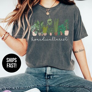 Op de afbeelding: Donkergrijs t-shirt met een afbeelding van verschillende potplanten en het witte woord "hoardiculturist". Het shirt heeft een ronde hals en korte mouwen. De stof lijkt zacht en comfortabel.