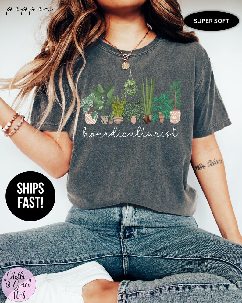 Op de afbeelding: Donkergrijs t-shirt met een afbeelding van verschillende potplanten en het witte woord "hoardiculturist". Het shirt heeft een ronde hals en korte mouwen. De stof lijkt zacht en comfortabel.