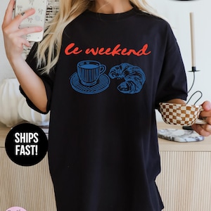 May include: Black oversized t-shirt with the phrase "Le Weekend" in red script. A blue illustration of a coffee cup and croissant is printed below the text. The shirt is labeled "Super Soft".