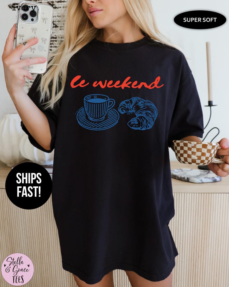 May include: Black oversized t-shirt with the phrase "Le Weekend" in red script. A blue illustration of a coffee cup and croissant is printed below the text. The shirt is labeled "Super Soft".