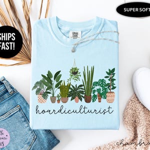 Op de afbeelding: Lichtblauw T-shirt met een afbeelding van diverse potplanten en het woord "hoardiculturist" in zwart schrift. Een zilveren ketting met een kleine hanger ligt op het shirt. Het shirt is voorzien van de labels "Super Soft" en "Comfort Colors".