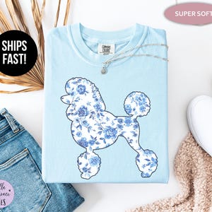 May include: Light blue t-shirt featuring a poodle silhouette filled with a blue floral pattern. The shirt has a crew neck and short sleeves. The text "SHIPS FAST!" is in a black circle. The text "SUPER SOFT" is in a pink oval.