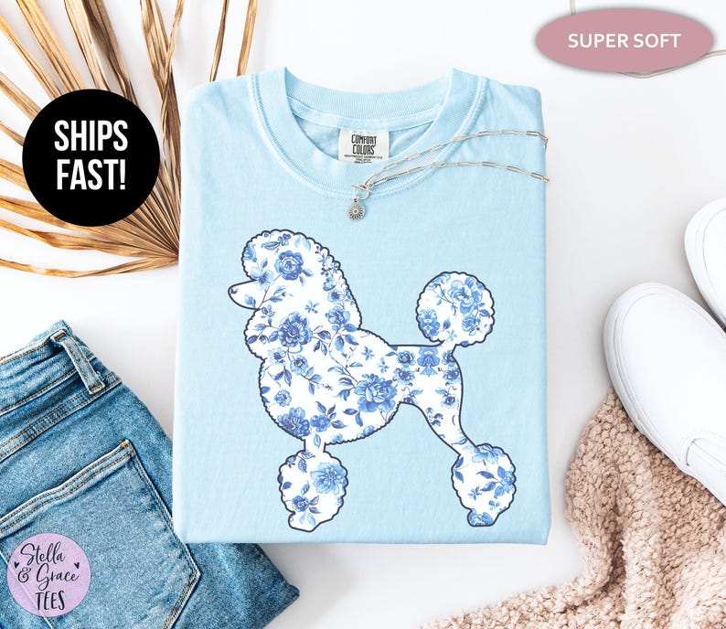 May include: Light blue t-shirt featuring a poodle silhouette filled with a blue floral pattern. The shirt has a crew neck and short sleeves. The text "SHIPS FAST!" is in a black circle. The text "SUPER SOFT" is in a pink oval.
