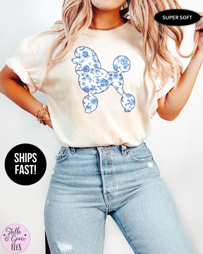 May include: Cream-colored t-shirt featuring a blue floral poodle design. The shirt has short sleeves and is paired with light-wash blue jeans. The text "SUPER SOFT" is visible. The brand name "Stella & Grace Tees" is also visible.