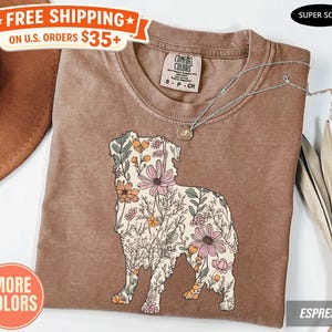 May include: A brown t-shirt with a floral dog design. The dog silhouette is filled with pink, orange, and cream-colored flowers and foliage. A silver necklace with a small pendant is draped on the shirt.